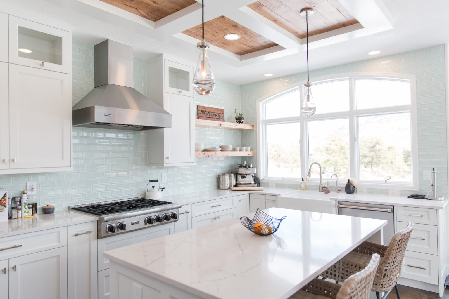 The view of a brightly lit, cool-toned renovated kitchen features a marble-topped kitchen island, new stove and hood, and windows looking out over local Colorado scenery.