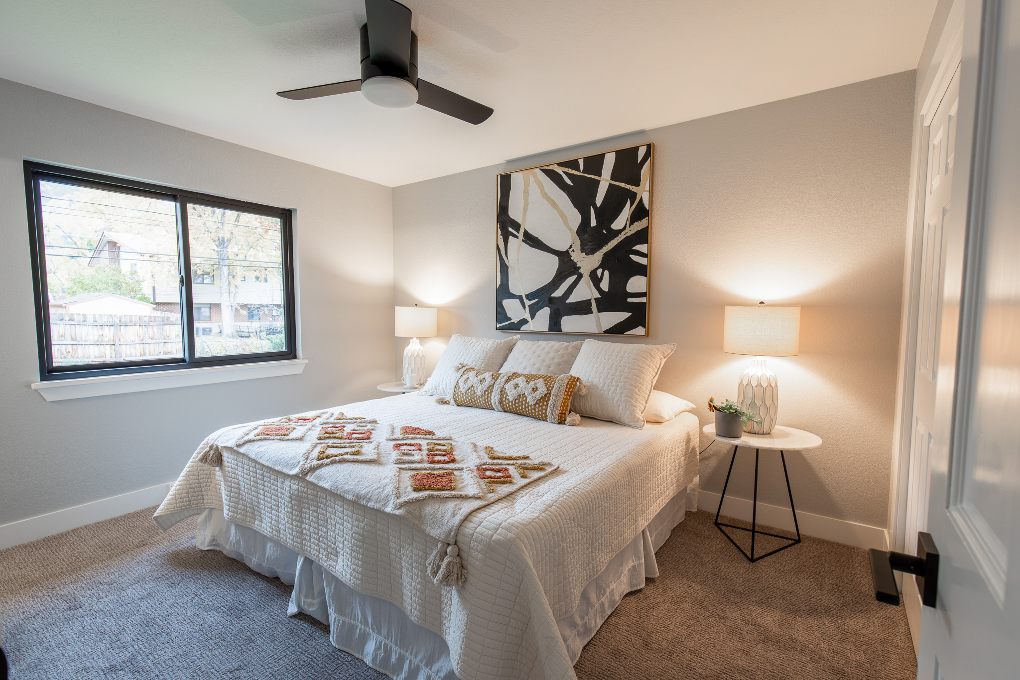 A furnished queen bed is at the center of a newly renovated bedroom, between two nightstands with lamps providing warm light throughout the room.