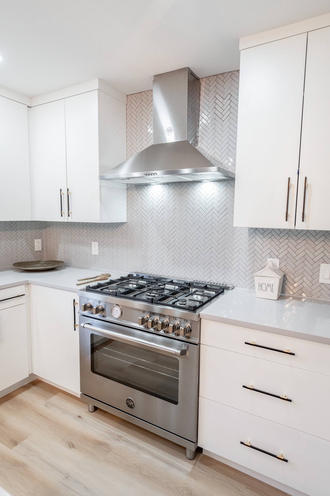 A new stainless steel oven, stove, and hood are set into white custom cabinetry. Against the wall, gray tile is positioned in a chevron pattern.
