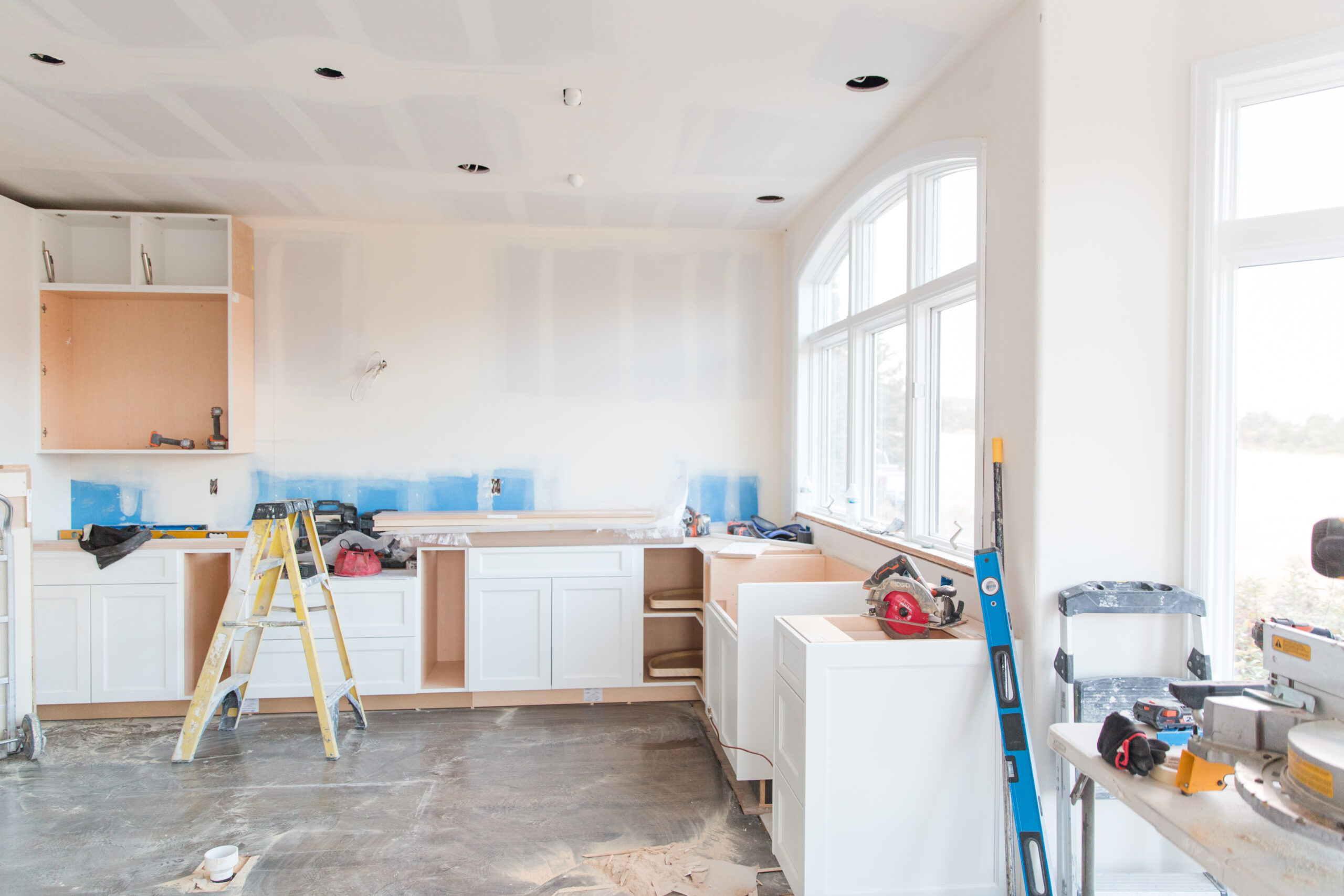 In a semi-remodeled kitchen, the cabinetry is being installed. The ceilings and walls are primed for paint, and the floor remains unfinished.