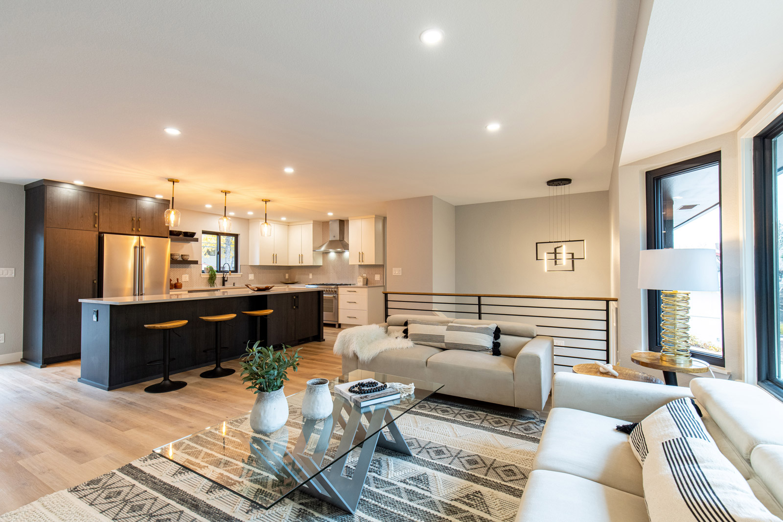 Two couches line the walls of a newly renovated living room. In the center of the room, a glass table sits atop a large decorative rug. In the background, a brightly lit kitchen.